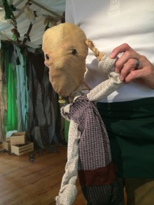 A puppet with a head that looks like a potato but is made of paper on top of a body made of cloth while a hand can be seen just behind the puppet's head