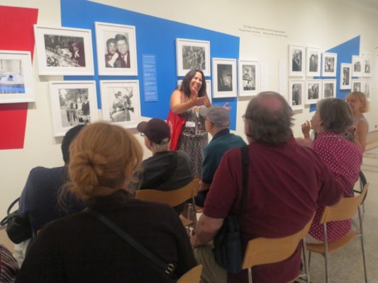 A group sits facing a wall of photos. A woman stands facing the group with her hands stretched out towards them.