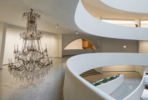 The right side of the image shows a side of the stacked spiral walkway of the Guggenheim. On the left side in a large chandelier extending from the ceiling to the floor of the walkway.