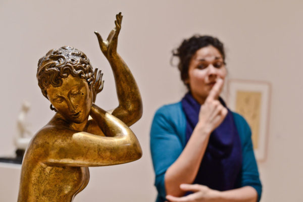 On the left side of the image is a bronze statue with both arms bent and raised. On the right side, set back and out of focus, is a woman signing, one arm raised to her face.