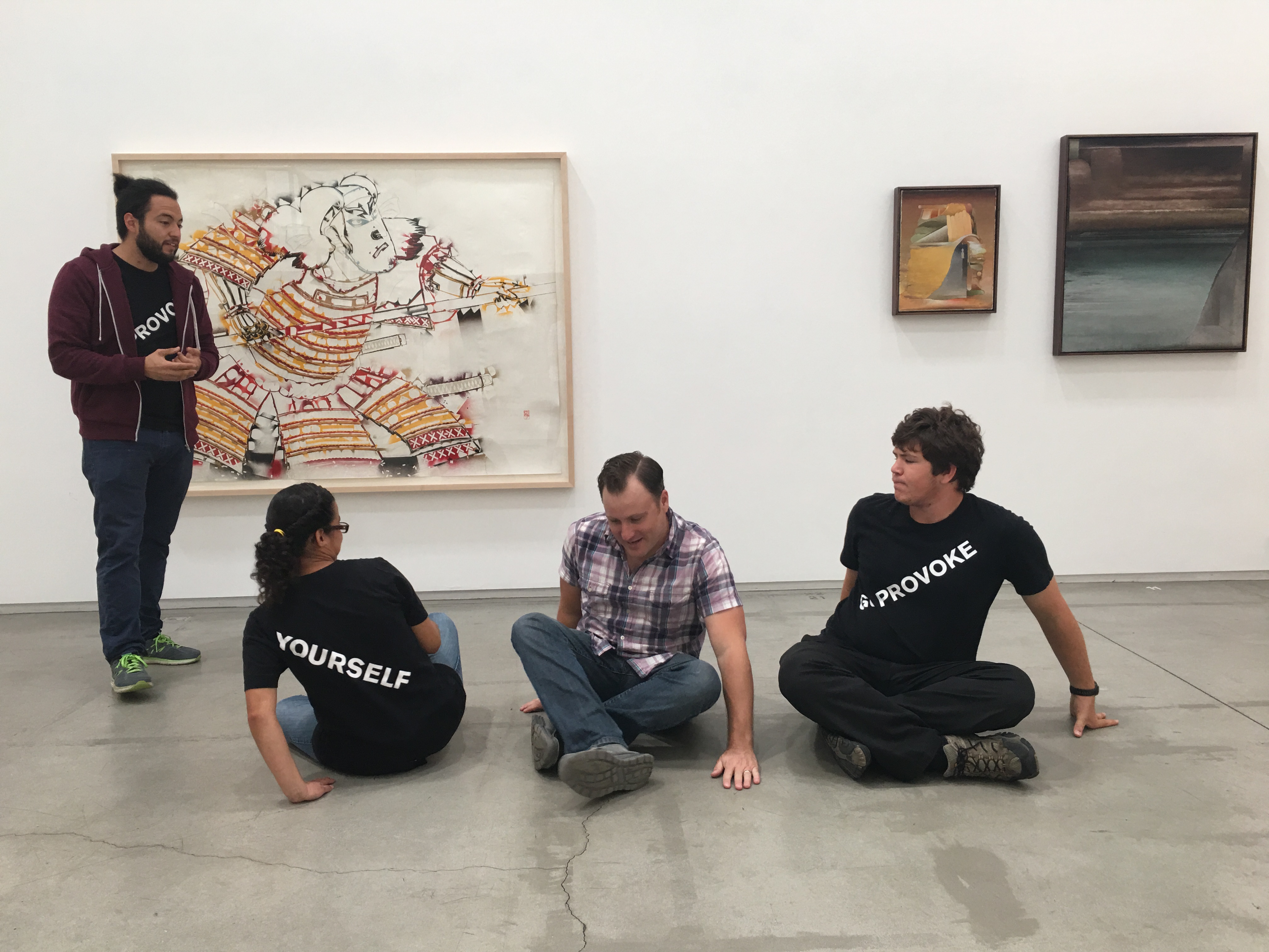 Four people in a white gallery with cement flooring. Three sit cross-legged facing various directions. One on the left stands looking towards the others. Behind them, three works of art hang.