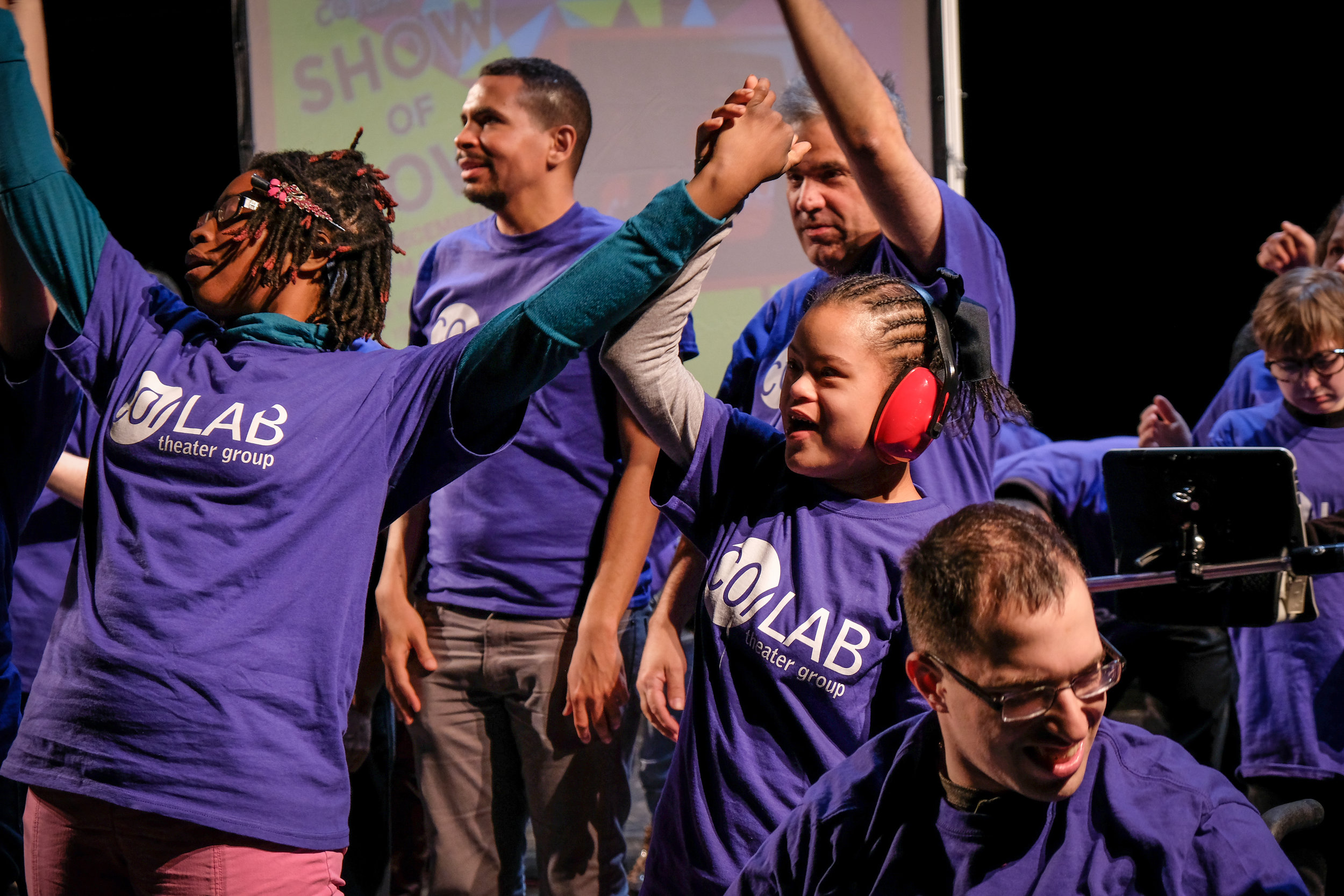 Seven actors of various ages wear purple CO/LAB shirts. Each is looking in a different direction, several have arms raised.