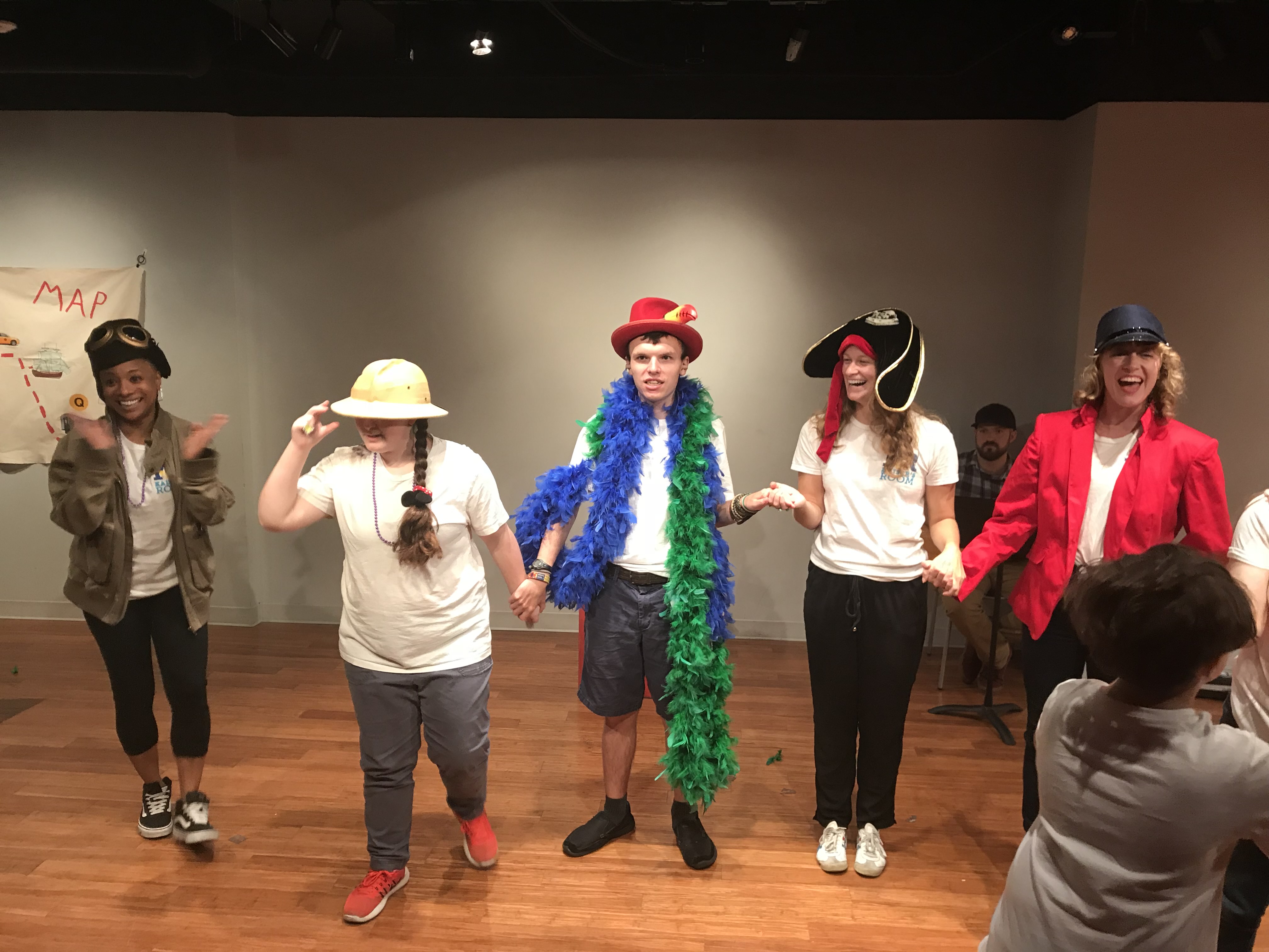 Five young people with various hats and feather boas hold hands and clap, smiling