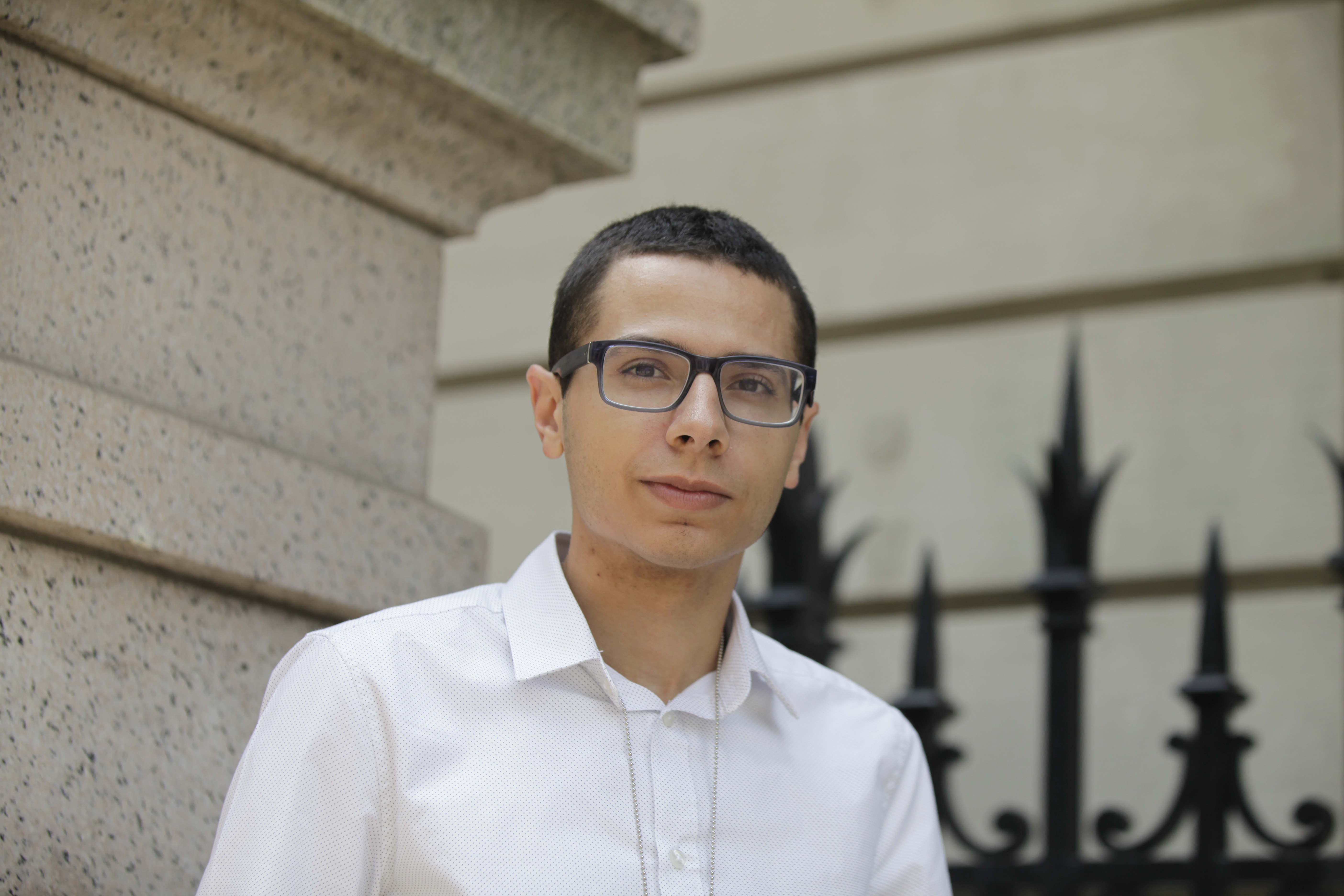 A young man with medium-dark skin wears glasses and stands in front of a light-grey stone building and iron fence.