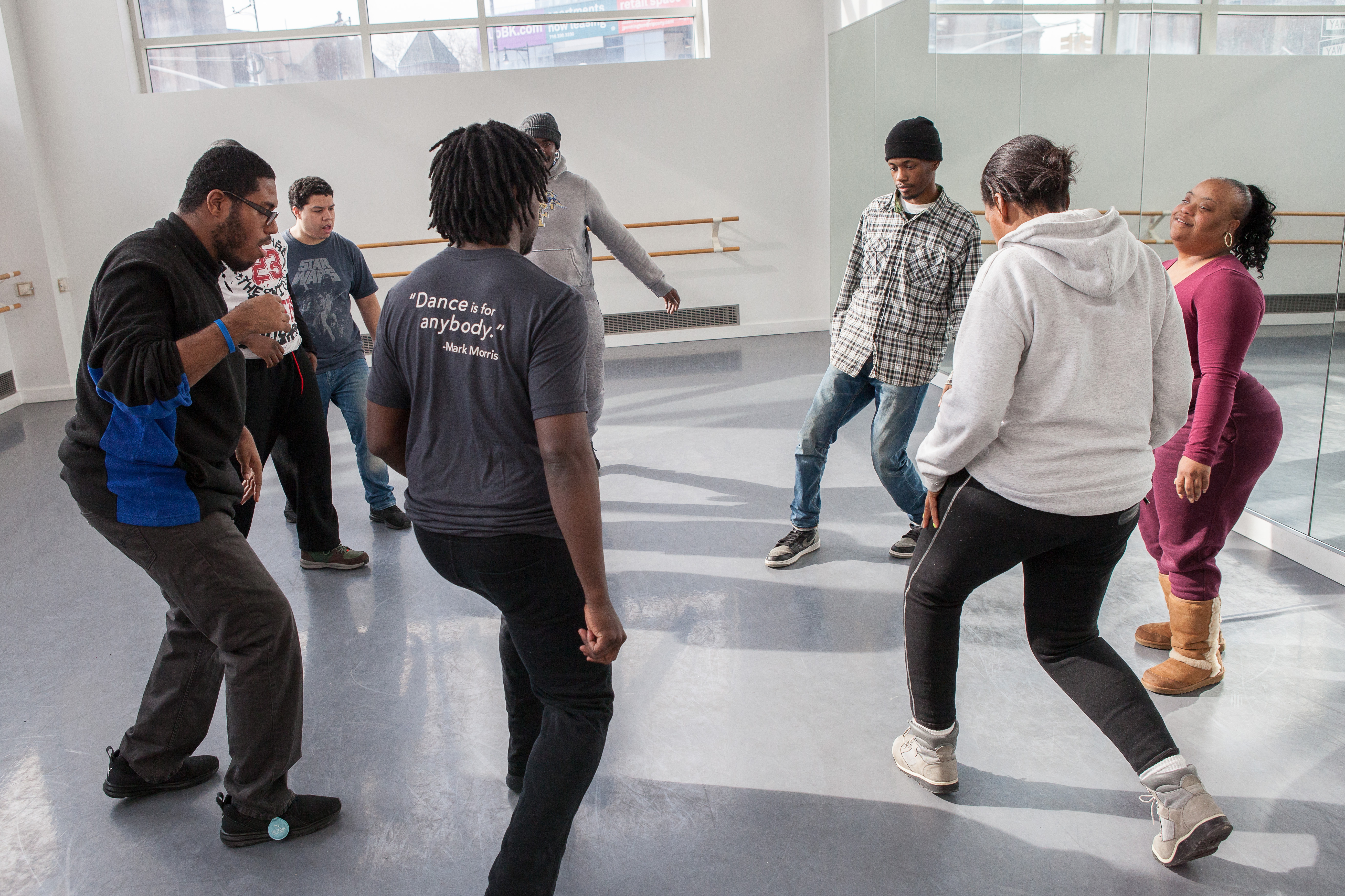 a group of six people of various ages stand in a circle in a brightly lit mirrored dance studio with legs bent and arms swinging in motion