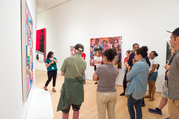 A group of visitors stand in a brightly lit gallery watching a gallery guide signing about a colorful painting