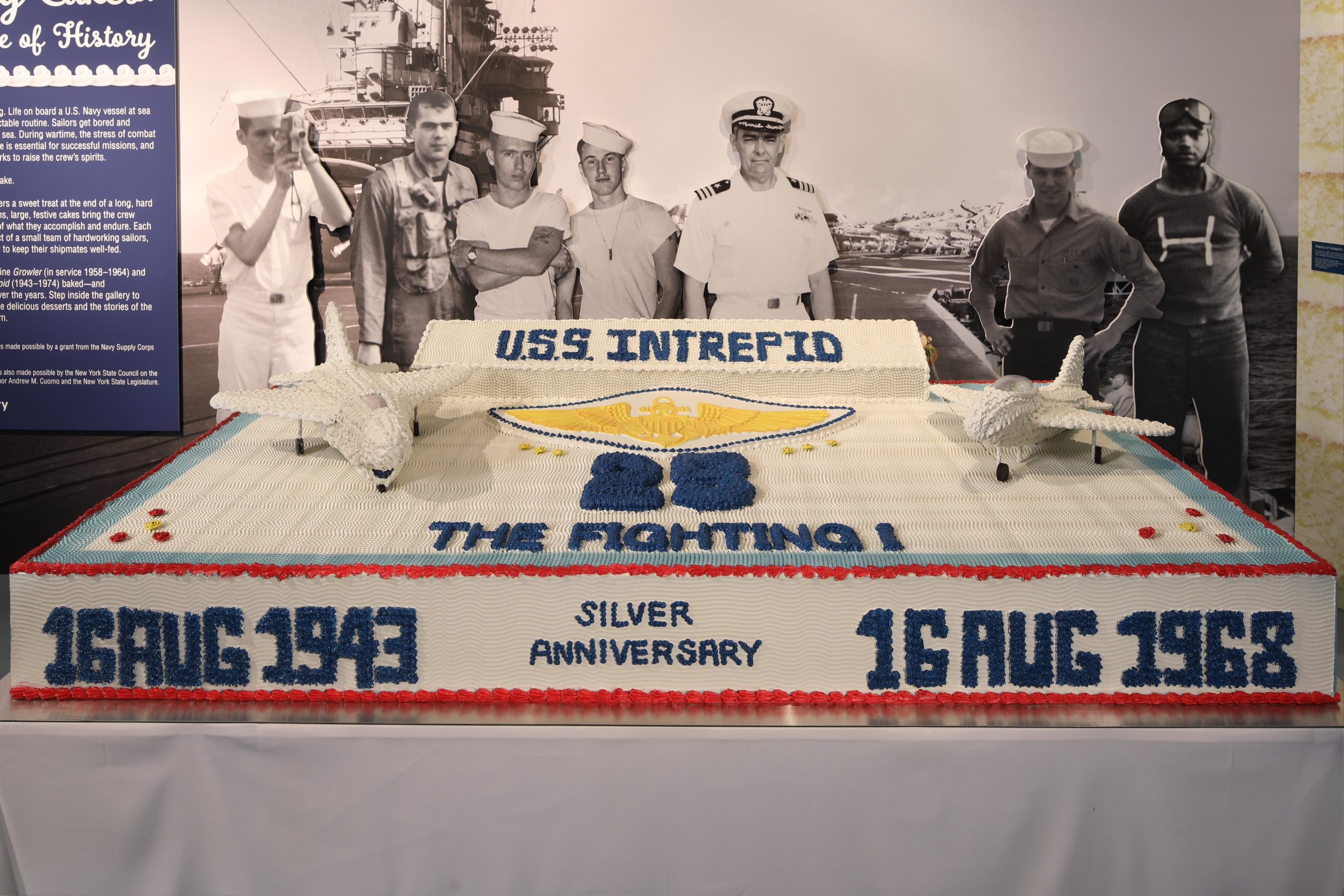Photo of full-size reproduction of Intrepid's giant 25th anniversary cake with life-size photos of crew members behind it