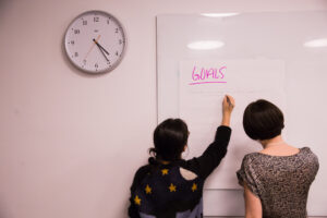 Two people with their backs to the camera write on a large piece of paper stuck to a whiteboard. The paper has the word "GOALS" written in large letters at the top of the page.