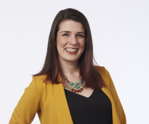 A smiling woman with light skin and shoulder length brown hair. She wears a mustard yellow blazer over a black V-neck shirt, along with a colorful beaded necklace.