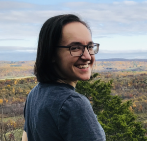 A smiling person with olive skin and shoulder length dark brown hair looks over their shoulder at the camera. They wear glasses with dark rectangular frames and a blue t-shirt. Behind them is an expanse of trees and hills beneath a cloudy sky.