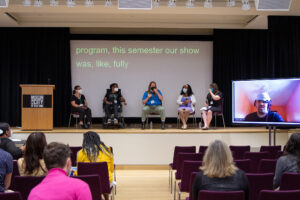 Five people sit on a stage giving a panel talk, with a sixth panelist present via Zoom on a large screen.