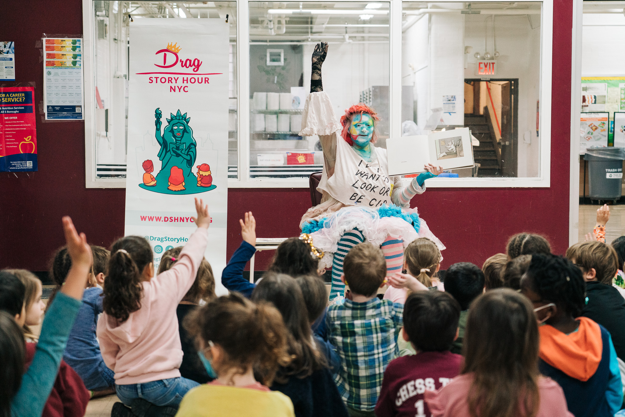 A drag artist with bright pink hair and a sunny blue sky with clouds painted across their face holds up a picture book to a room full of children seated on the floor.