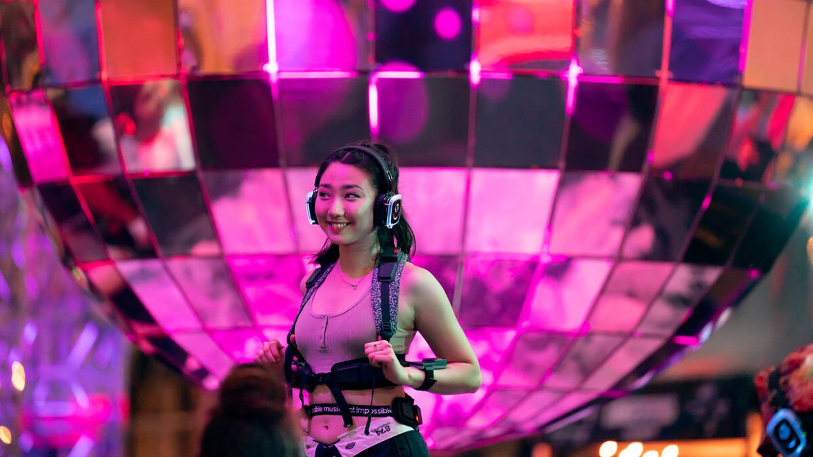 A person smiles, bathed in purple light, as they stand in front of a massive glittering disco ball while wearing over-ear headphones.